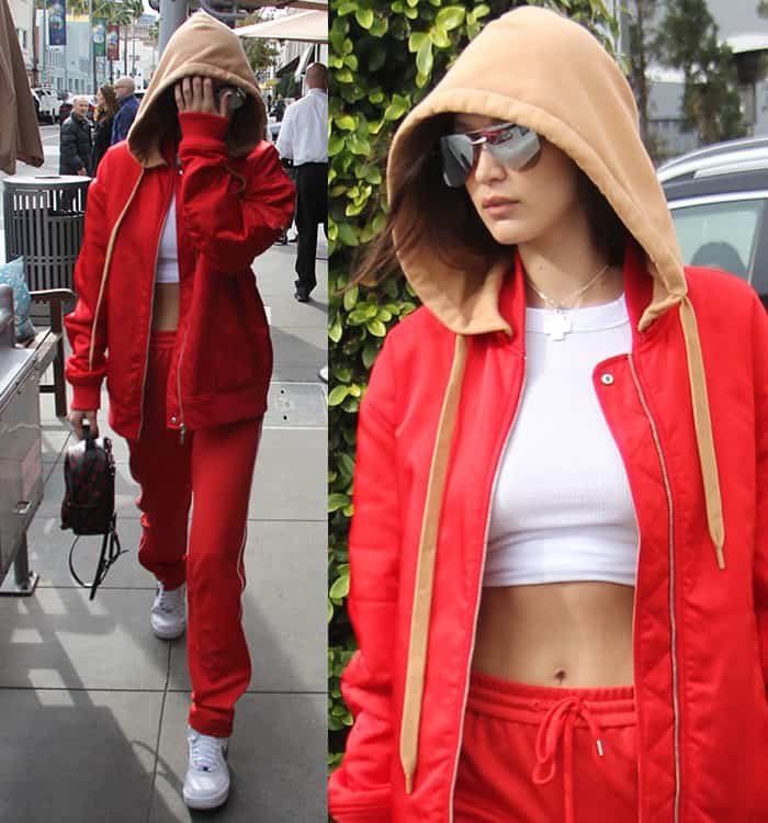 Bella Hadid steps out at Il Pastaio in a bold red tracksuit layered with a tan-hooded jacket, pairing it with a white crop top, Nike Air Force 1 sneakers, Quay Playa sunglasses, and a Louis Vuitton Palm Springs Mini Backpack