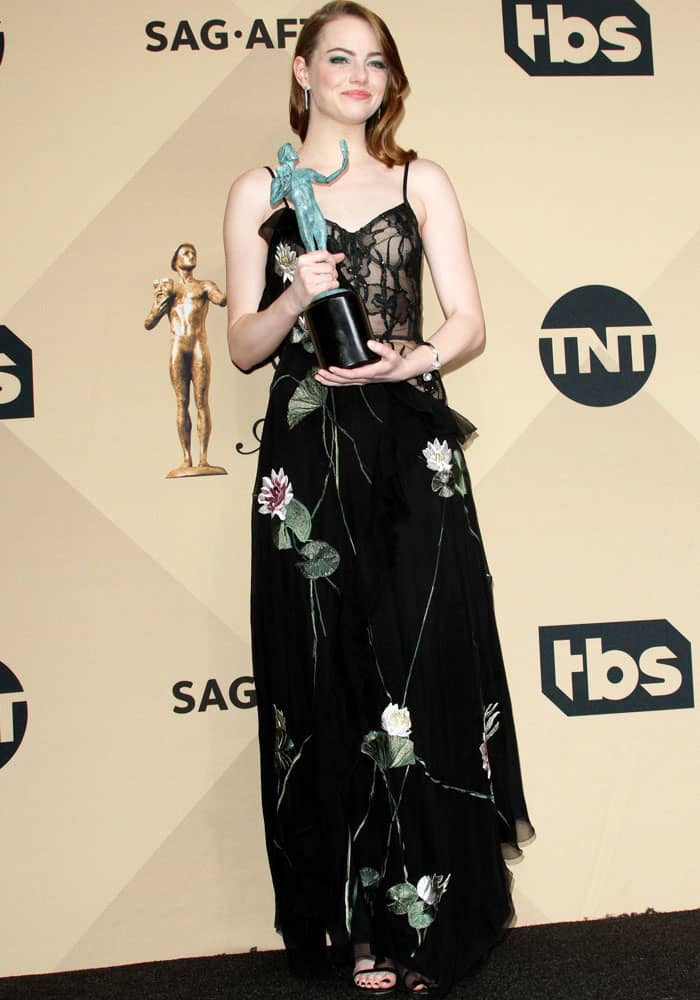 Donning an elegant black Alexander McQueen dress that combined the sophistication of a three-fourths embroidered gown with a hint of allure, featuring a surprise sheer boudoir corset top, Emma Stone shows off her trophy in the press room at the SAG Awards