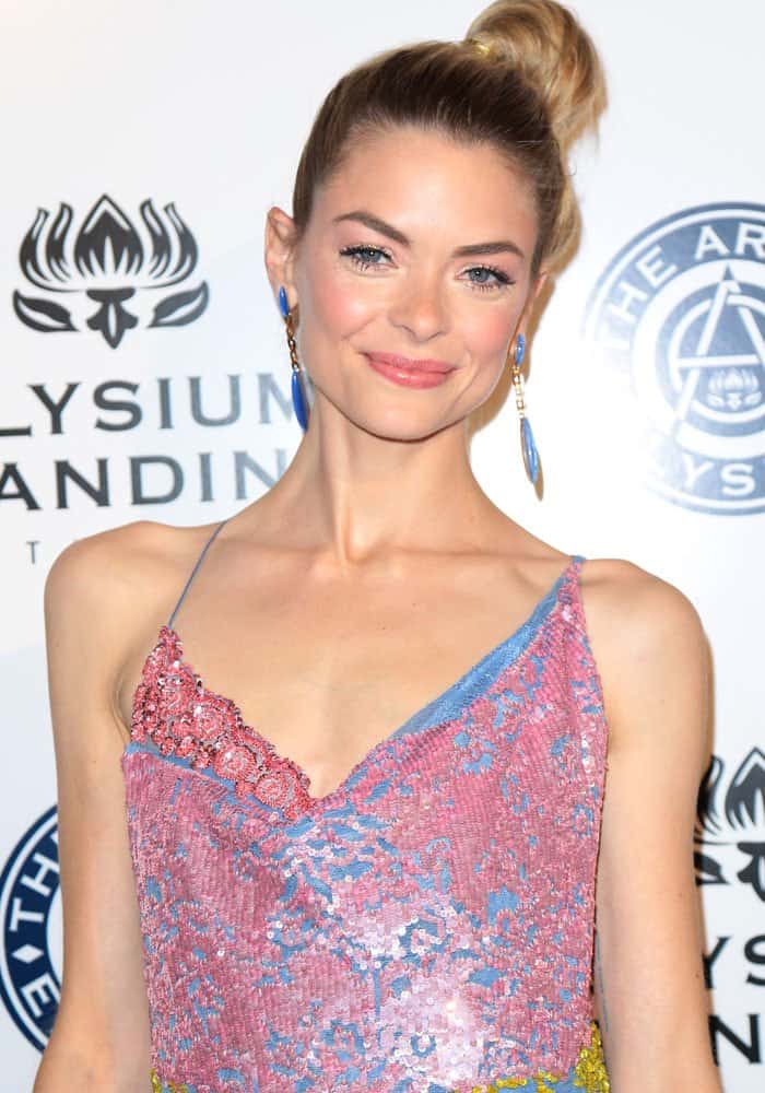 Jaime King at the Art of Elysium gala with her hair styled in a sleek topknot, showing off her statement blue drop earrings and soft, natural makeup that complements her vibrant sequined dress