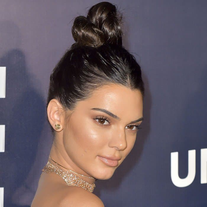 Kendall Jenner sports a sleek top knot that highlights her flawless makeup and glowing skin, while her understated gold stud earrings add a subtle touch of elegance to the overall look