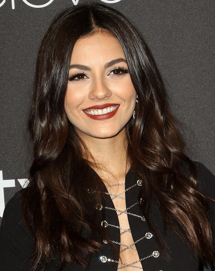 Victoria Justice completes her sultry look with voluminous brunette waves, bold red lipstick, and dramatic lashes, perfectly complementing her chain-laced gown