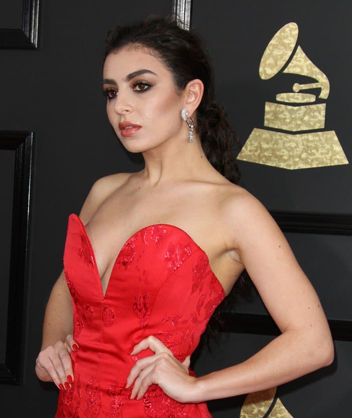 Charli XCX styles her hair in a voluminous, textured ponytail, flaunts a bold red manicure, and strikes a confident pose to complement her glamorous red carpet look at the 2017 Grammy Awards