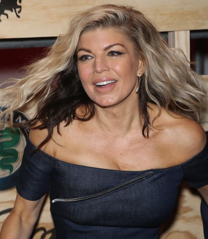 Fergie styled her voluminous, wavy blonde hair with a dark ombré touch, complementing her radiant, natural makeup look featuring soft pink lips and a glowing complexion