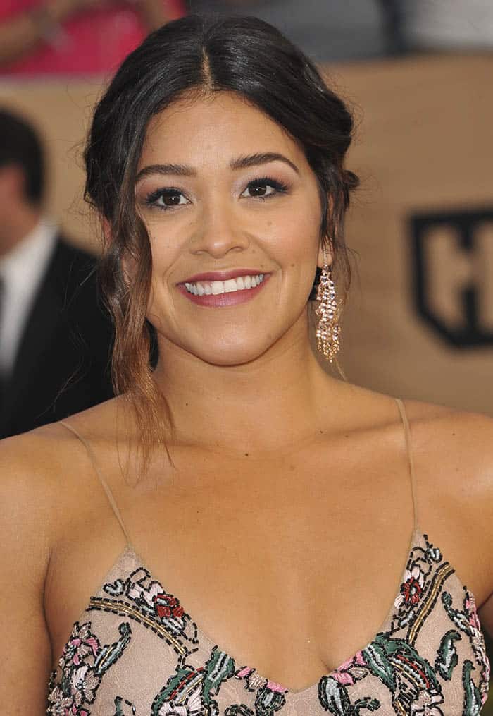 Gina Rodriguez radiates charm with her romantic updo featuring loose waves, soft neutral makeup, and sparkling chandelier earrings that perfectly complement her intricately embroidered gown