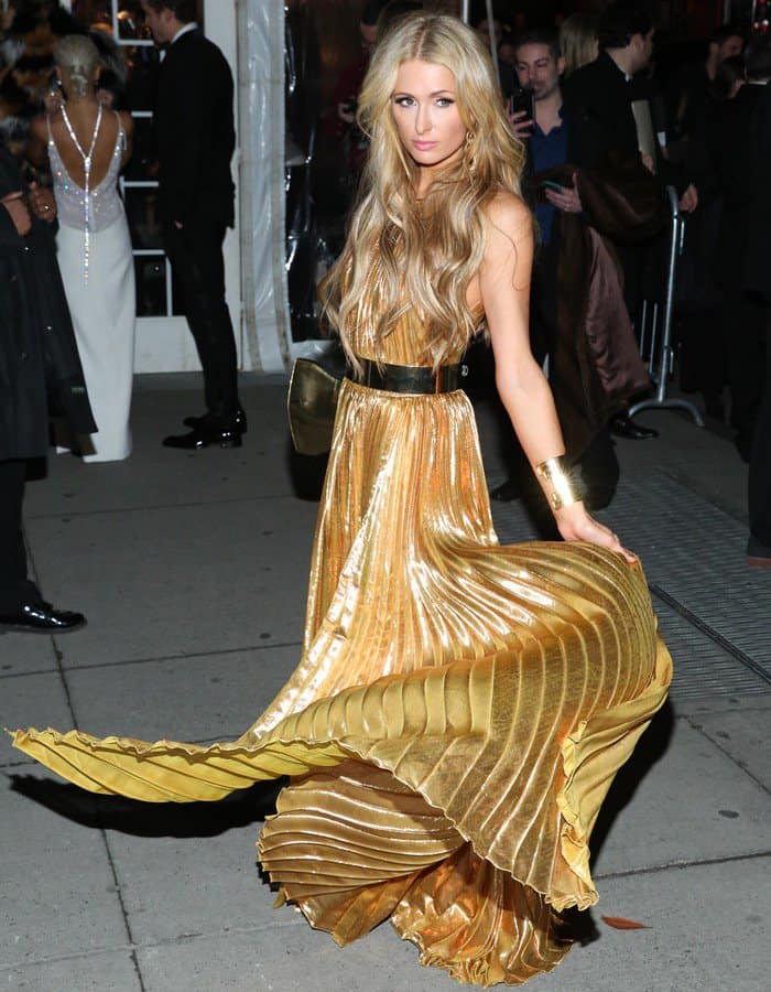 Paris Hilton demonstrates how to wear metallic gold with confidence, twirling in a pleated Maria Lucia Hohan gown that showcases its dramatic flow and elegant shimmer—perfectly accessorized with bold gold cuffs and a black belt for a modern touch