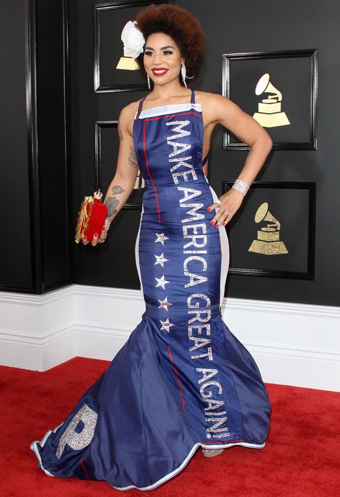 To replicate Joy Villa's bold look, opt for a custom-designed mermaid gown with political or statement text in glittery embellishments, pair it with a red heart-shaped clutch, sparkling platform heels, and finish with a white floral hair accessory and bold red lipstick