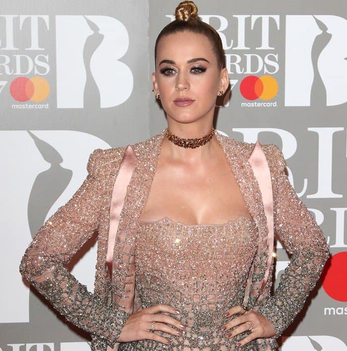 Katy Perry captivates with a sleek top knot hairstyle and bold eye makeup, complemented by a shimmering pink ensemble