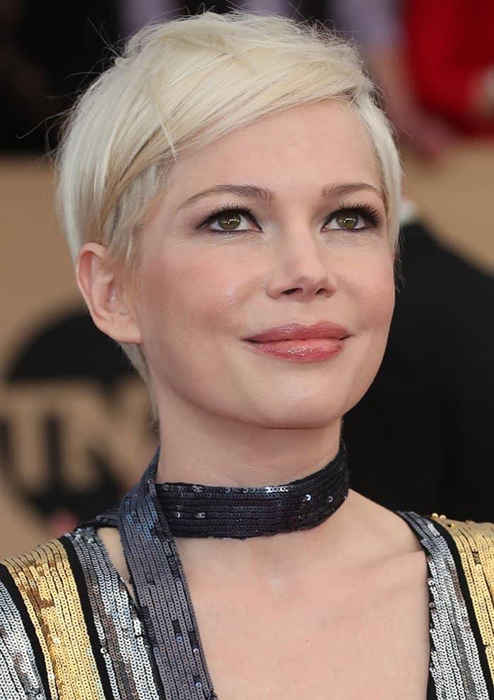 Michelle Williams exudes modern elegance with her platinum pixie cut, subtle smoky eyes, glossy nude lips, and a shimmering sequined choker that perfectly complements her futuristic gown