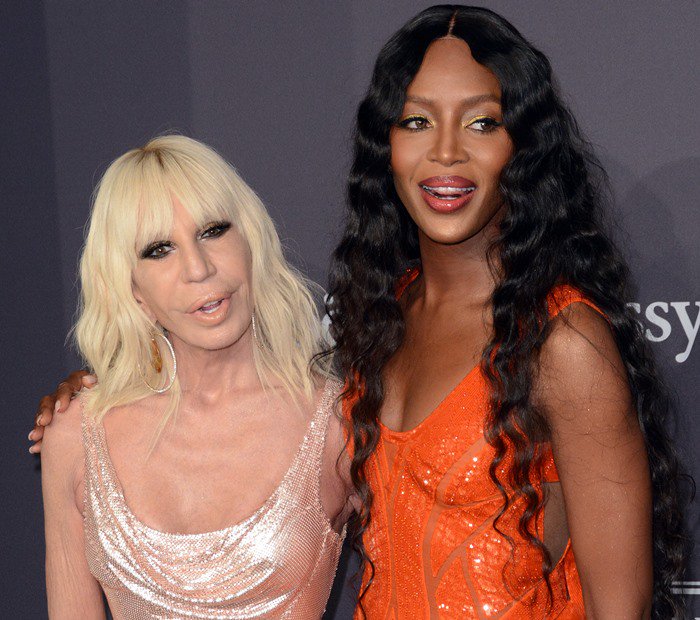 Naomi Campbell stuns in a glowing orange Atelier Versace gown with flawless makeup, standing beside Donatella Versace, who dazzles in a shimmering blush pink dress