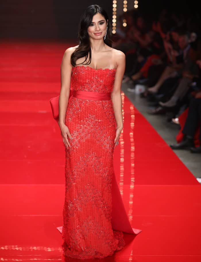 Diane Guerrero dazzles in a strapless red gown adorned with intricate silver embellishments and a bold satin bow, exuding timeless elegance on the runway
