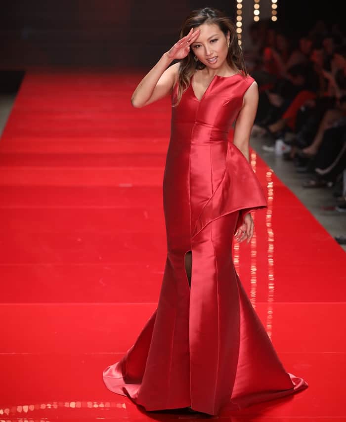 Jeannie Mai radiates confidence in a sleek red satin gown with asymmetrical peplum detailing and a thigh-high slit, perfectly complementing her playful runway salute