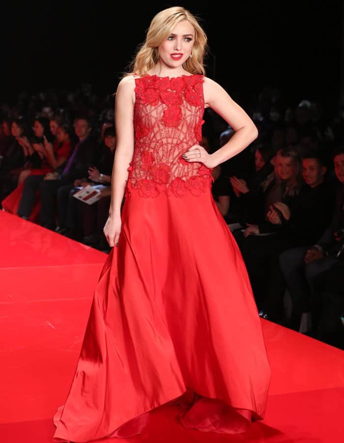 Peyton List stuns in a bold red gown featuring a floral-embellished sheer bodice and flowing satin skirt, exuding glamour and sophistication on the runway