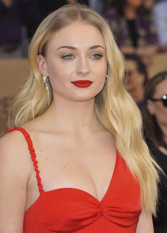 Sophie Turner captivates with her classic Hollywood waves, bold red lips, and diamond drop earrings, perfectly complementing her striking red gown for an effortlessly glamorous look