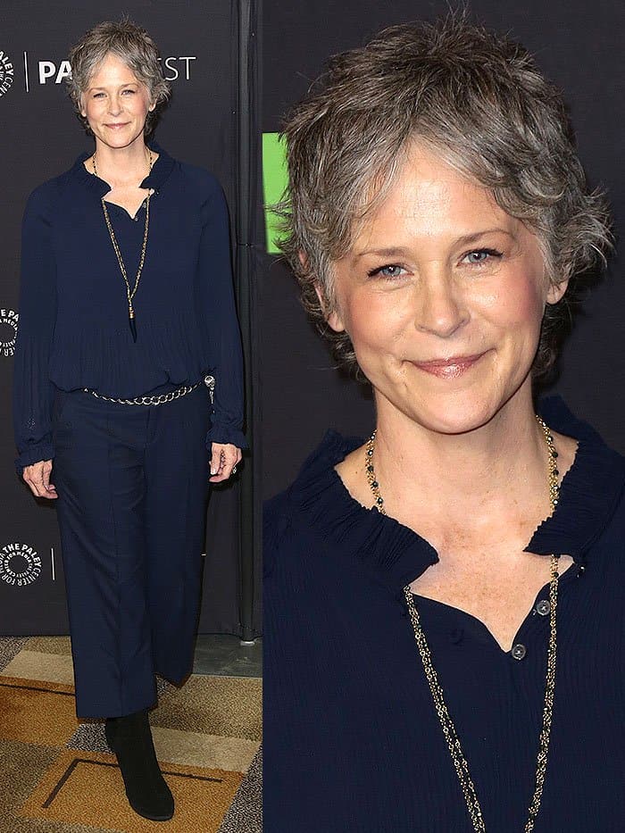 Melissa McBride exudes effortless sophistication in a navy ruffled blouse, matching cropped trousers, and timeless black suede boots, accessorized with a delicate chain belt
