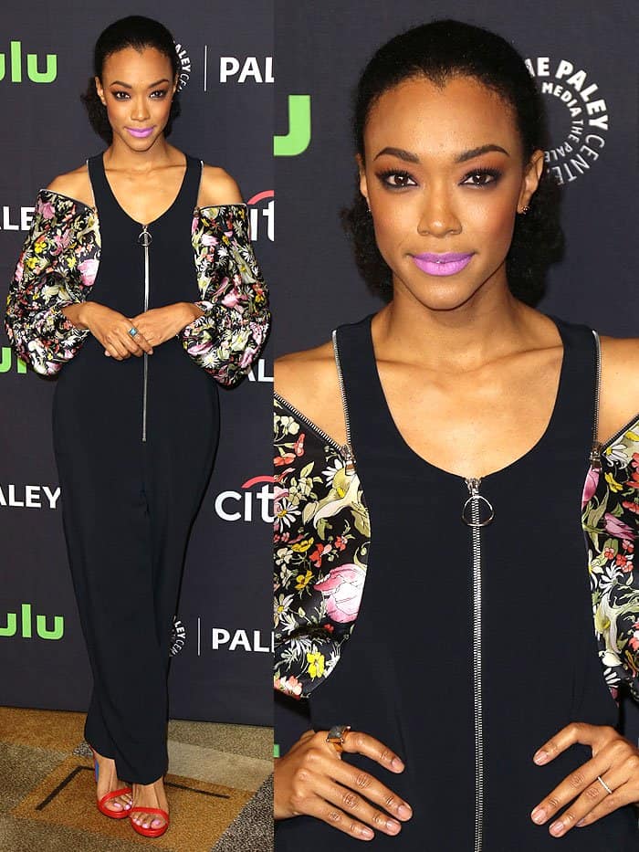 Sonequa Martin-Green makes a bold statement in a navy zip-front jumpsuit with floral satin ruched sleeves, vibrant purple lipstick, and eye-catching red ankle-strap sandals with blue accents