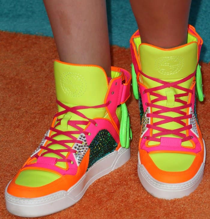 JoJo Siwa keeps it signature bold with eye-catching neon high-top sneakers featuring vibrant orange, pink, and lime green panels, sparkling embellishments, and playful side pouches for an extra dose of fun