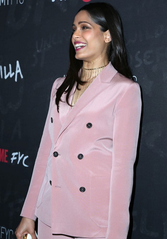 Freida Pinto wears a pink double-breasted pantsuit paired with a gold I.V.I. necklace that does not quite complement the sleek lines of her outfit