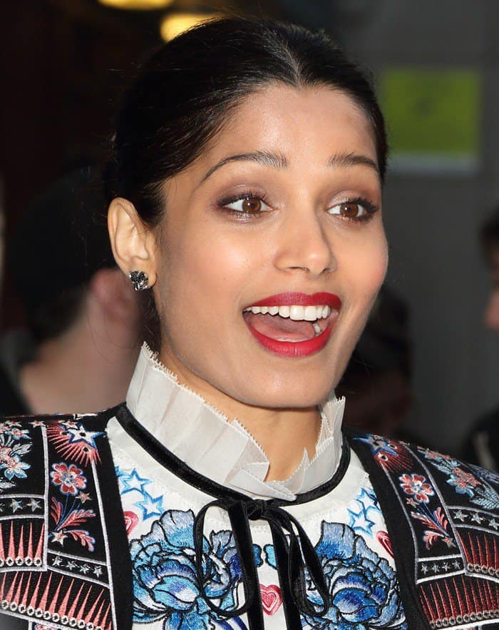 Freida Pinto wears elegant black stud earrings, a bold red lip, and her hair slicked back into a chic, polished updo