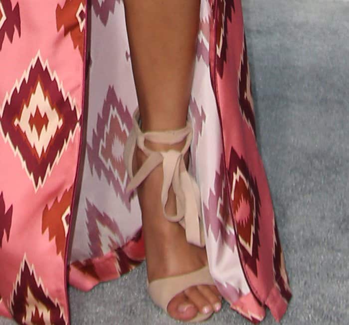Jhene Aiko showcases her elegant feet in nude Schutz sandals, tied delicately around her ankles, paired with a fresh, glossy nude pedicure that complements her patterned pink-and-maroon robe dress