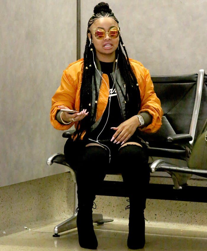 Blac Chyna nailed airport chic with an edgy mix of ripped black leggings, knee-high lace-up boots, an oversized orange bomber jacket, and statement sunglasses—perfect for effortless, stylish travel