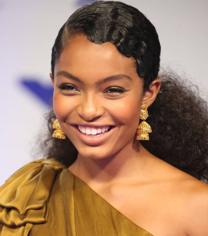 Yara Shahidi’s curls are styled in a chic side-parted ponytail, perfectly framing her face and highlighting her radiant smile and bold, gold statement earrings