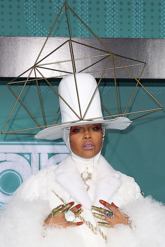Erykah Badu stuns in a surreal ensemble featuring a towering gold pyramid hat, dramatic white fur accents, and bold metallic accessories