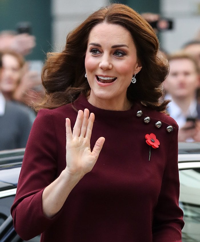Kate Middleton greets the crowd with a warm wave, radiating elegance in her burgundy tunic dress adorned with metallic button details and a Remembrance poppy pin
