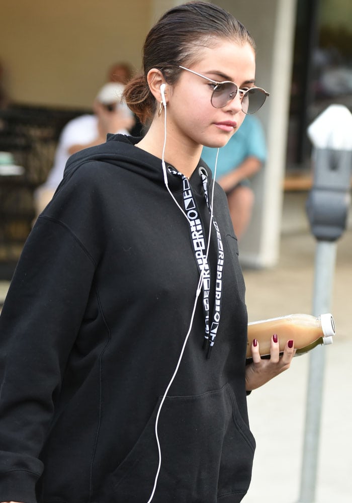 Selena Gomez steps out in a casual black hoodie, holding a juice bottle and listening to music, showcasing her laid-back street style