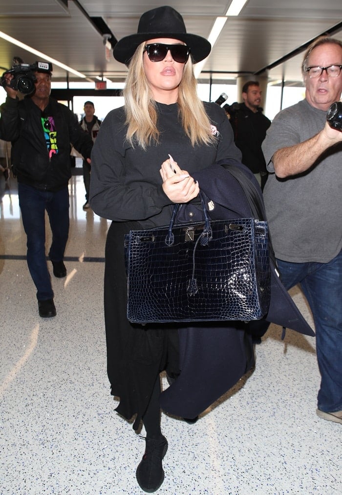 Khloe Kardashian sported a black pull-over sweatshirt, a wide-brim hat, black workout leggings, one more sweatshirt tied around her waist, designer sunglasses, and a long black trench coat