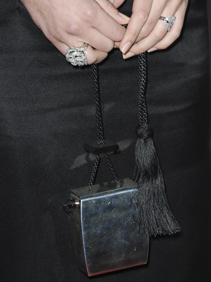 Margot Robbie carries a sleek, reflective silver clutch by THE ROW, featuring a unique sculptural shape, black cord handle, and a bold tassel accent for a modern, minimalist finish