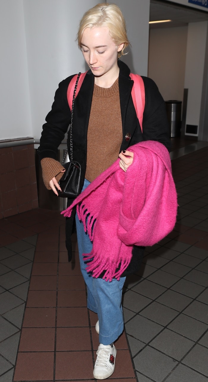 Saoirse Ronan wearing wide-legged jeans styled with a brown knitted sweater, a black woolen coat, an oversized pink scarf, and white sneakers