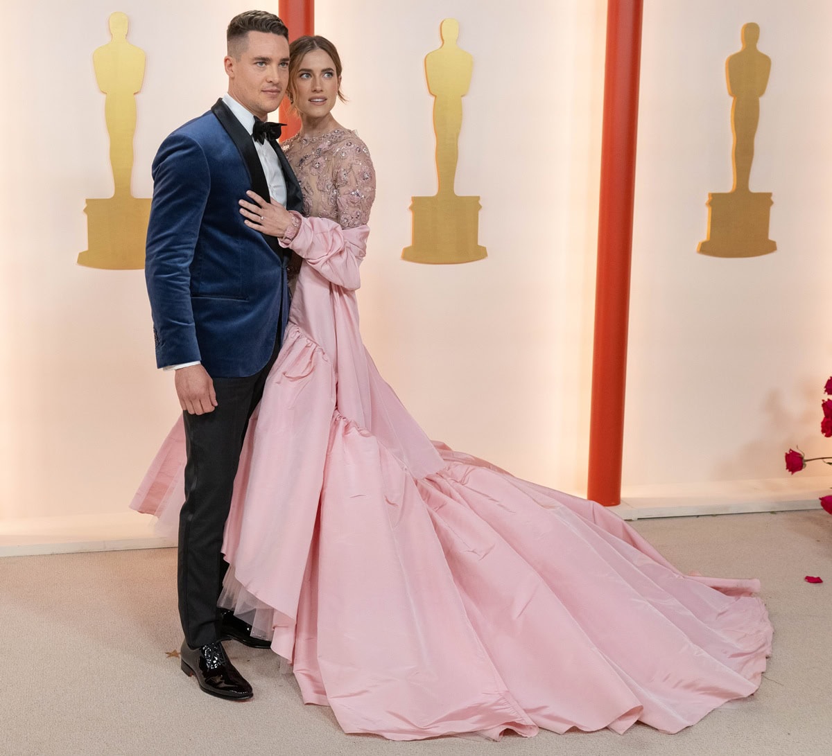 Allison Williams turned heads at the 95th Academy Awards in a dramatic pink-and-silver Giambattista Valli haute couture gown with floral embellishments, a feather hem, and an extravagant cape train, joined on the red carpet by fiancé Alexander Dreymon
