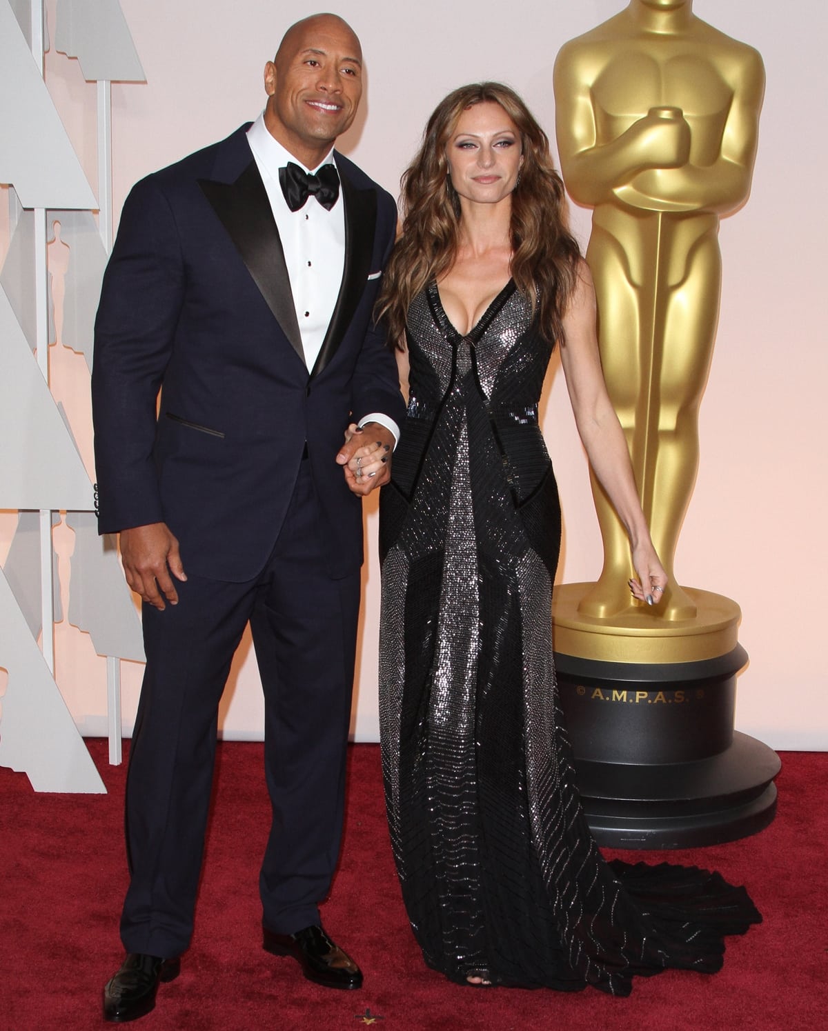 Dwayne Johnson looks dapper in a classic navy tuxedo, while Lauren Hashian stuns in a shimmering black gown with metallic accents, making a glamorous appearance at the Academy Awards