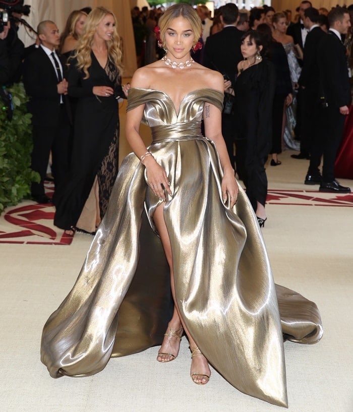 Jasmine Sanders commands attention in a show-stopping metallic gold gown with a dramatic thigh-high slit, paired perfectly with strappy gold sandals and bold floral hair accents