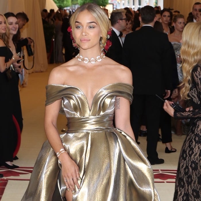 Jasmine Sanders stuns in a custom metallic gold off-the-shoulder gown, paired with an elegant braided updo adorned with red roses and complemented by a statement pearl choker and bracelet at the 2018 Met Gala
