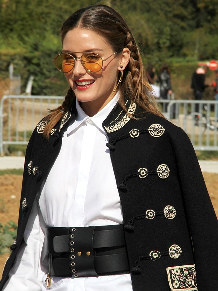 Olivia Palermo over-accessorizing with yellow-tinted sunglasses, dangling earrings, a gold-embroidered military jacket, a saddle belt, and braids in her hair.