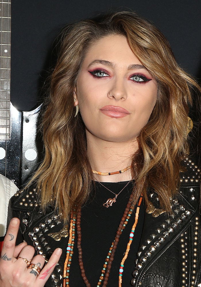 Paris Jackson rocks bold red-winged eyeliner, a nose ring, bohemian bead necklaces, and intricate finger tattoos as she strikes a devil horn pose in her studded leather jacket