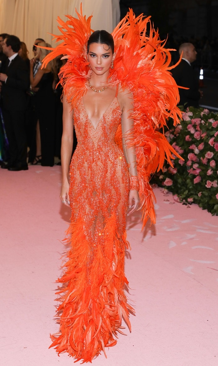 Kendall Jenner lit up the pink carpet in a sheer, figure-hugging Versace gown drenched in orange crystals and dramatic feathers, channeling Cher-inspired glam at the 2019 Met Gala