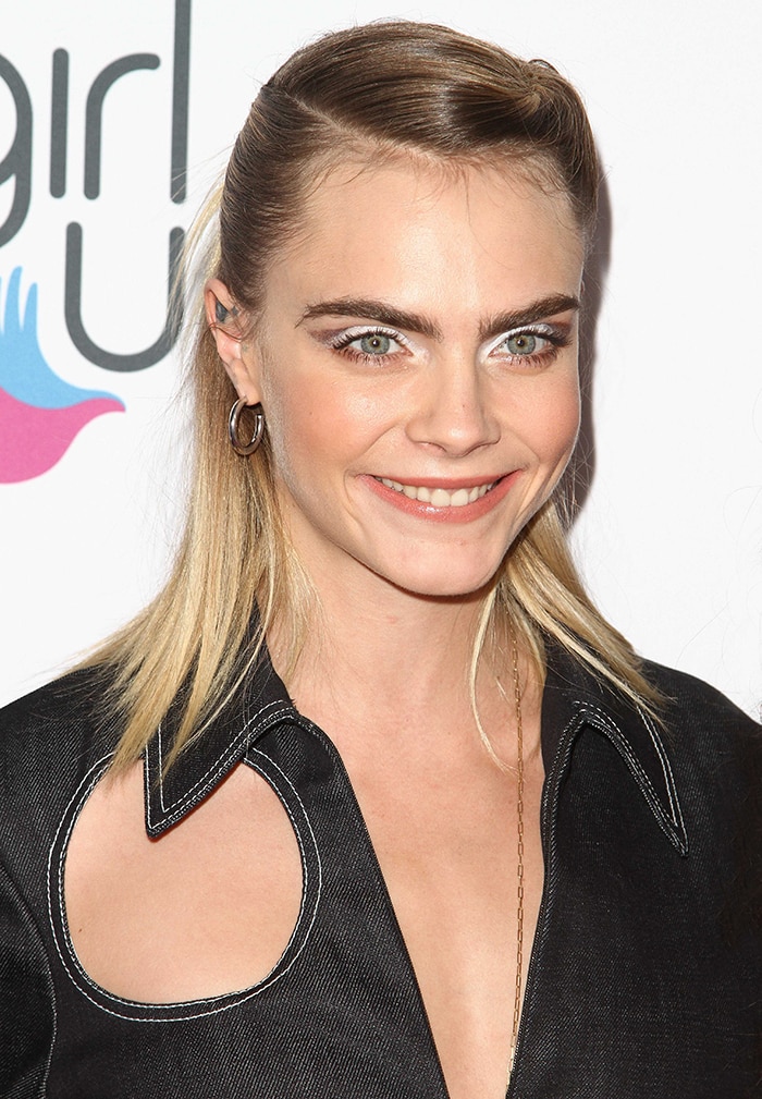 Cara Delevingne wears her shoulder-length blonde hair in a twisted half-up half-down style and sports a sweep of silver eyeshadow and peach lip shade