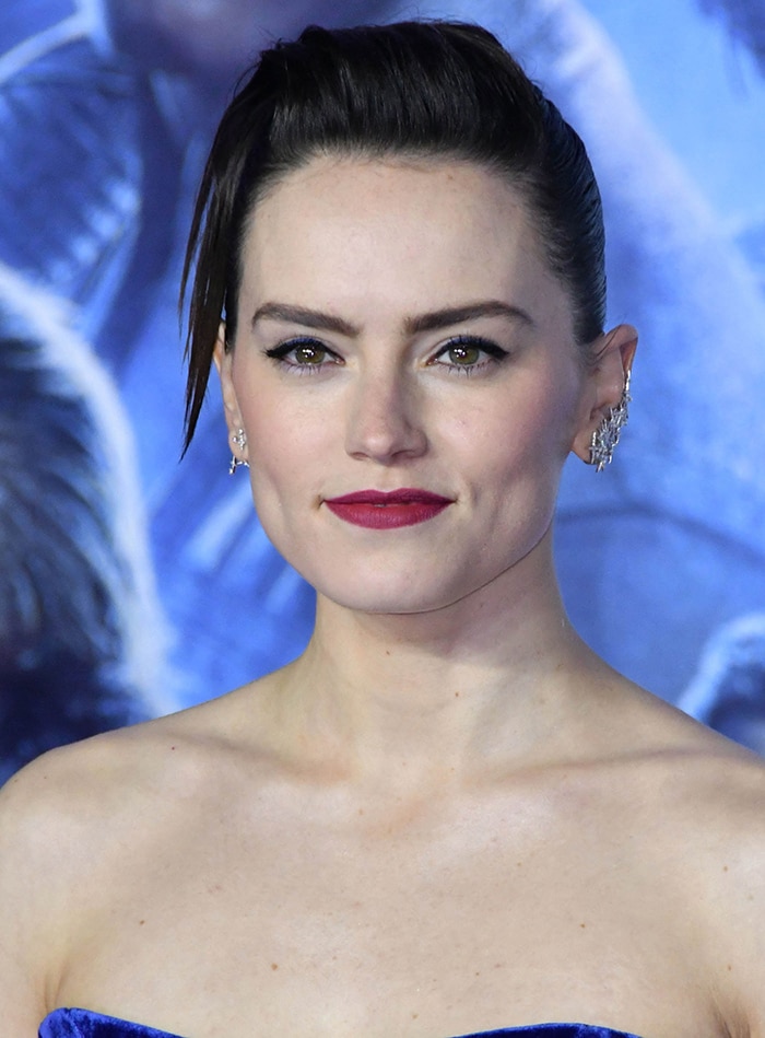 Daisy Ridley wears a slicked back hairstyle with blue tinge winged eyeliner and plum lipstick