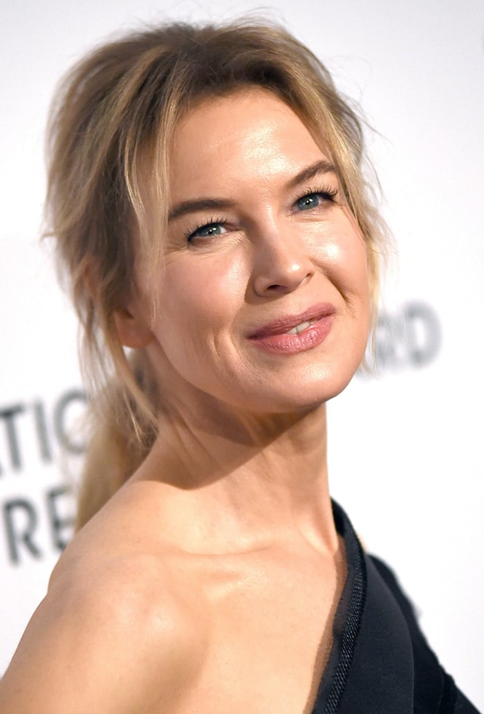 Renee Zellweger wears a messy ponytail with mascara, eyeliner, and pink lipstick