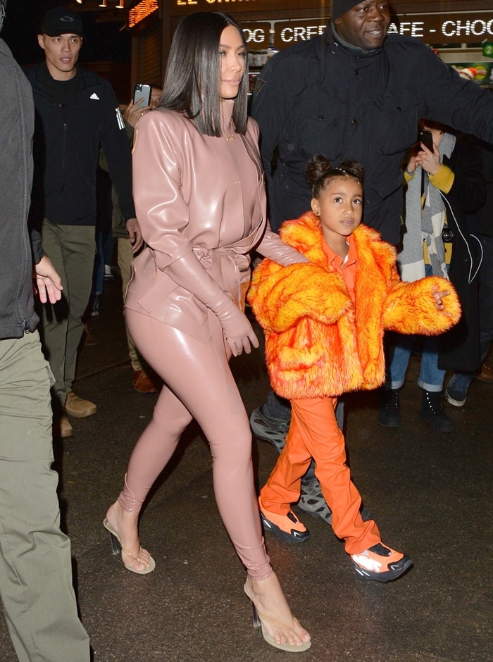 Kim Kardashian stuns in a pale-pink Balmain latex outfit paired with Yeezy Season 8 thong sandals, while North West brightens the scene in an orange fluffy coat and matching sneakers