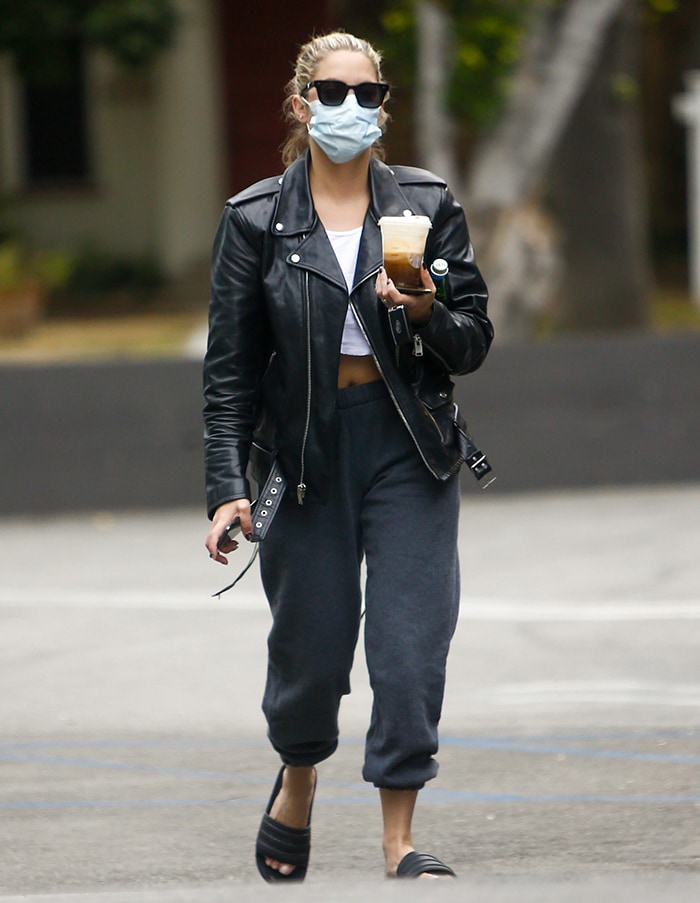 Ashley Benson keeps it casual yet edgy in a black leather biker jacket, white crop top, gray joggers, and Adidas slides, completing the look with sunglasses and a face mask during her solo coffee run