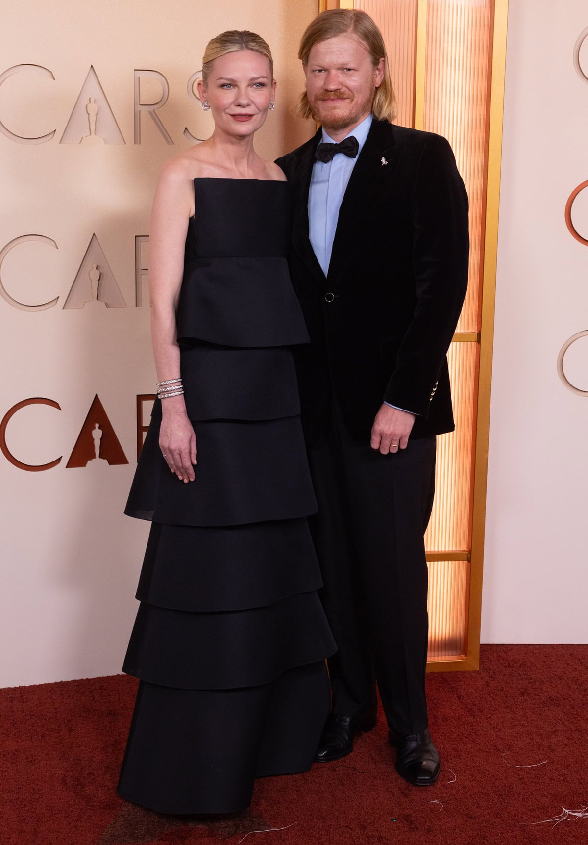 Kirsten Dunst stuns in a custom tiered Celine haute couture gown with a sharp square neckline, while Jesse Plemons complements her in a perfectly tailored black Lardini tuxedo, creating a sleek, architectural “power couple” silhouette at the Oscars