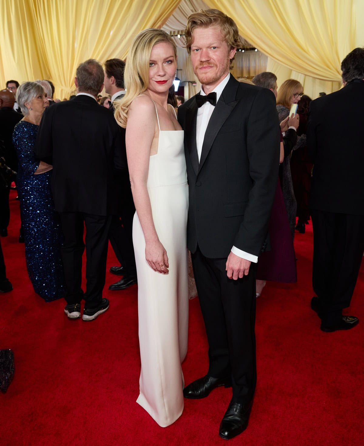Kirsten Dunst wears a sleek white Gucci column gown with delicate straps and a square neckline, while Jesse Plemons pairs a classic black tuxedo with polished patent shoes, channeling timeless red-carpet sophistication at the 96th Oscars