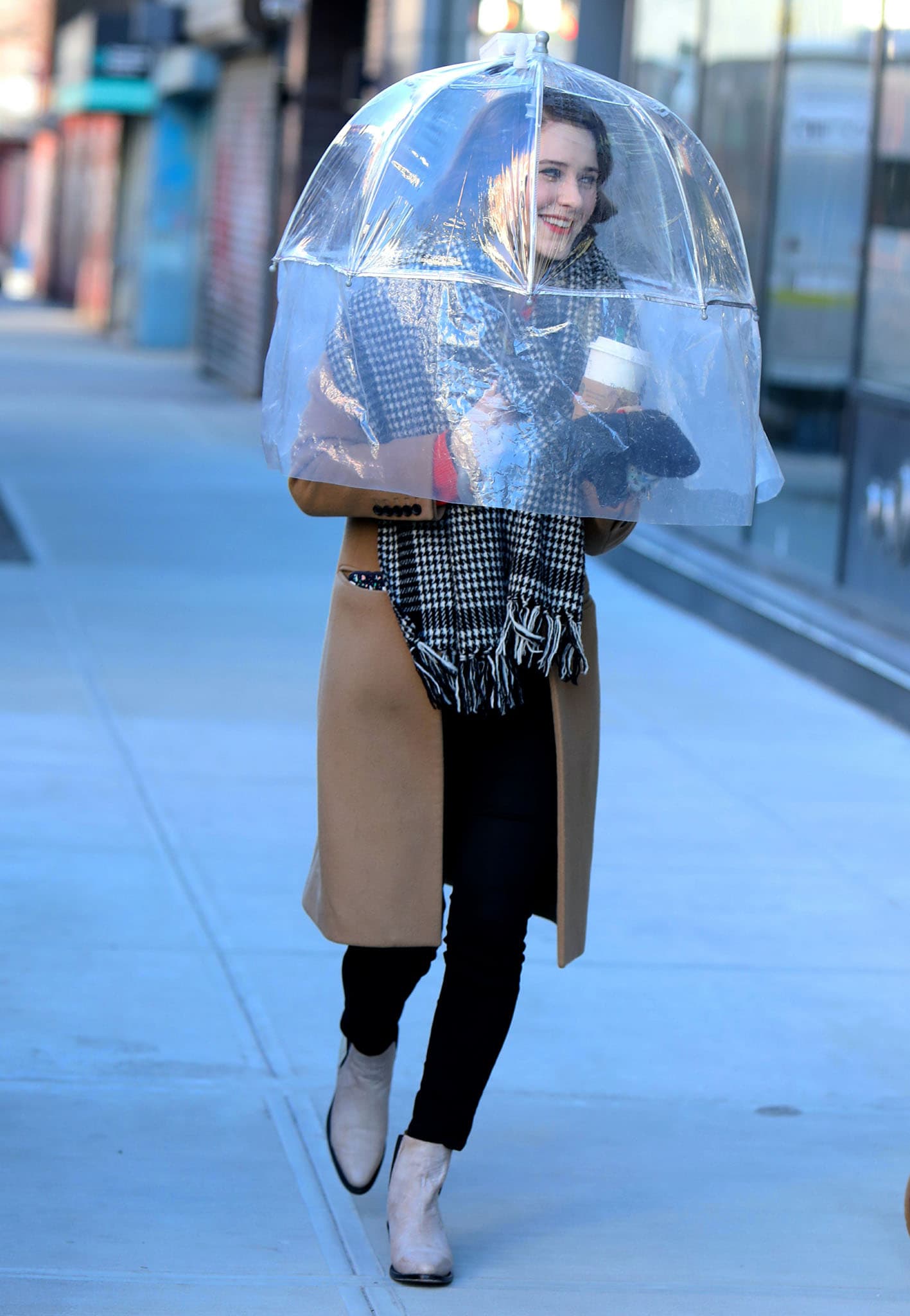 Rachel Brosnahan opts for a more relaxed look in black pants, tan coat, and checkered scarf