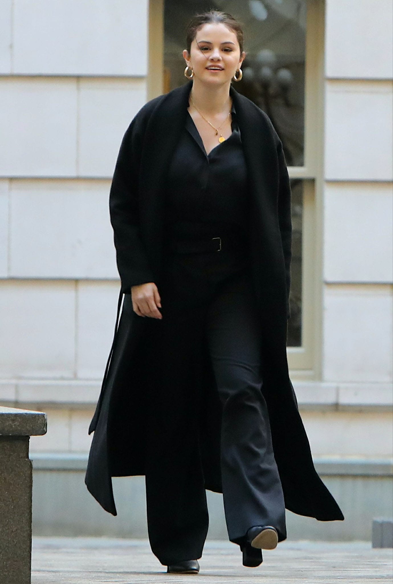 Selena Gomez wears a black coat over a black blouse tucked into a pair of high-waisted trousers