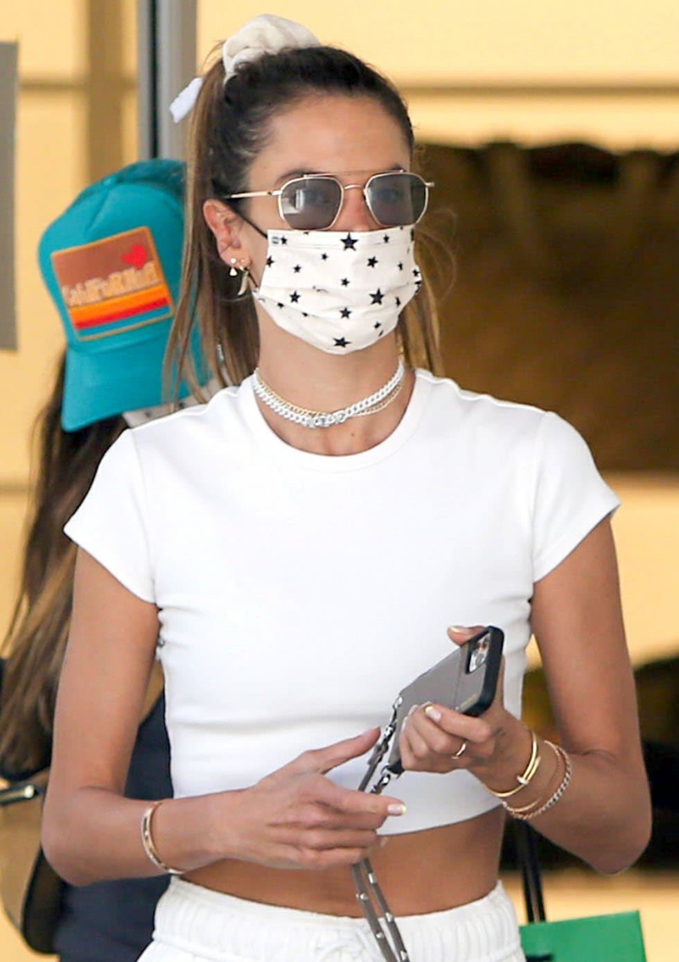 Alessandra Ambrosio looks chic with a high ponytail tied with a scrunchie, layered necklaces, and a star-patterned face mask