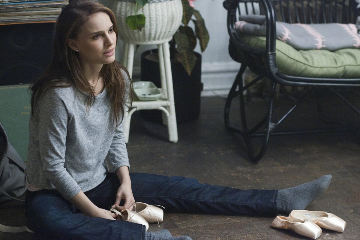Natalie Portman, as Nina Sayers in Black Swan, sits on the studio floor clutching her satin pointe shoes — a quiet moment that captures the film’s blend of vulnerability, obsession, and the disciplined beauty of ballet footwear