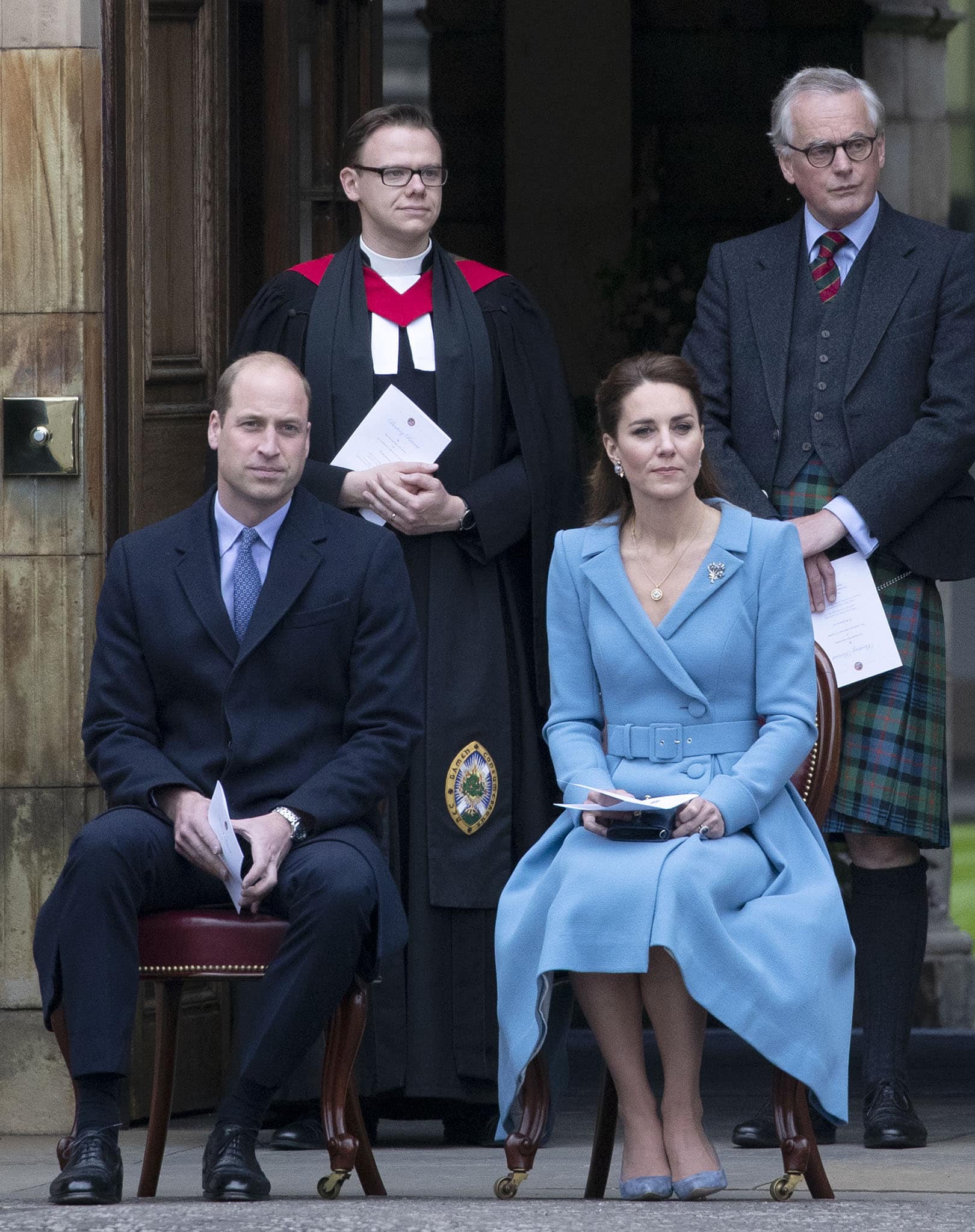 Prince William wears a navy blue suit with a printed necktie, while Kate Middleton opts for a light blue dress